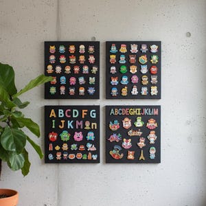 May include: Four black square art pieces are mounted on a gray concrete wall. Each piece features colorful, cartoon-like characters. Two pieces display the alphabet, while the others showcase a variety of figures. A potted plant is visible on the left.