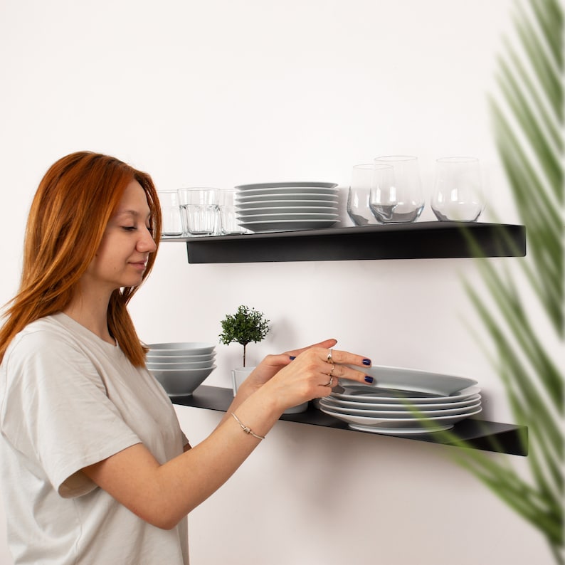 May include: A woman arranging gray plates on a black floating shelf. The shelf also holds clear glasses, bowls, and a small potted plant. The shelves are mounted on a white wall.