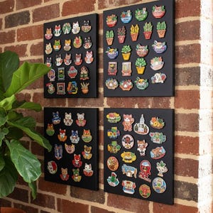 May include: Four black display boards mounted on a brick wall, each covered with colorful enamel pins. The pins feature cat and plant designs, showcasing a variety of colors and styles. The boards are arranged in a square formation.