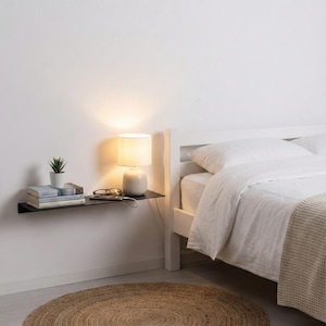 May include: A modern bedroom scene featuring a black floating shelf with a small potted plant, books, and a charging phone. A cream-colored lamp sits on the shelf, illuminating the items. A white bed with white linens and a woven rug complete the minimalist decor.