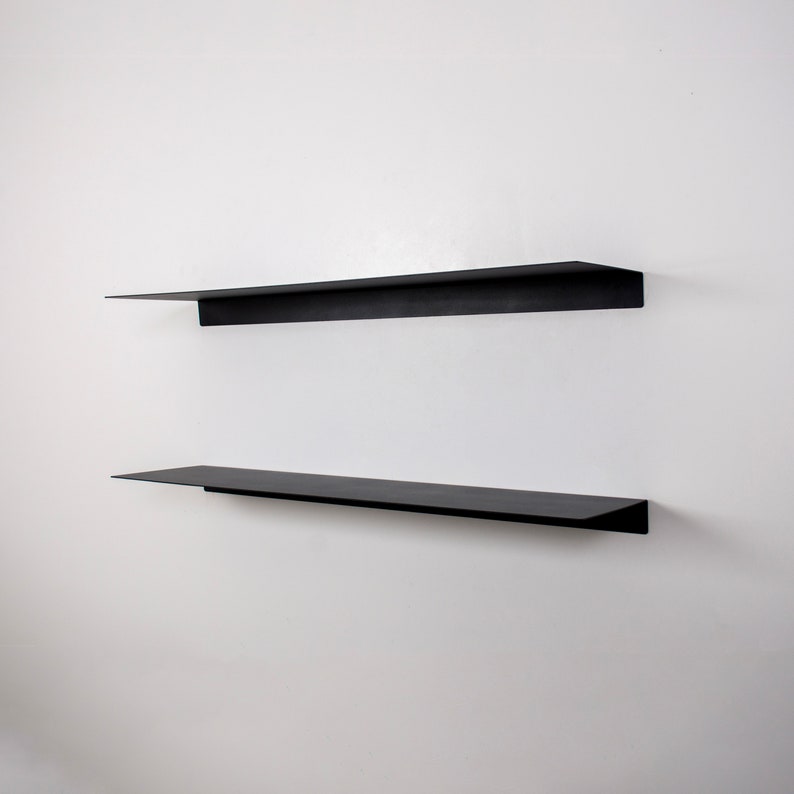 May include: Two black floating shelves mounted on a white wall. The shelves are rectangular with a minimalist design, perfect for displaying decor or storing items. The shelves are made of a matte black material.
