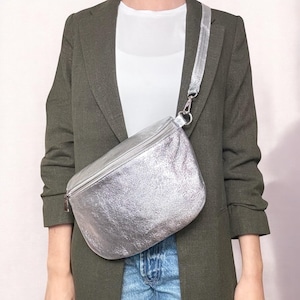 May include: A silver leather fanny pack with a silver zipper and a silver adjustable strap. The fanny pack is worn over a person's shoulder.