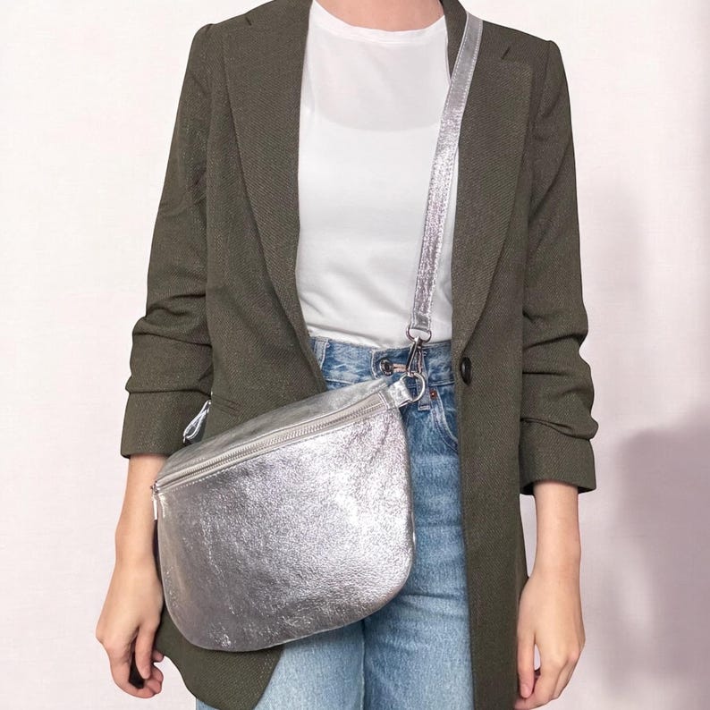 May include: A silver leather fanny pack with a silver chain strap. The fanny pack is worn over a green blazer and blue jeans.