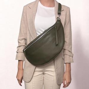 May include: A dark green leather fanny pack with a silver zipper and a long strap. The bag is worn across the body of a person wearing a beige blazer.