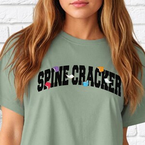 May include: A sage green t-shirt with the text "SPINE CRACKER" in black letters. The letters are decorated with small, colourful shapes.