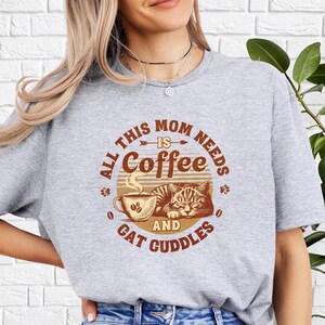 May include: Gray t-shirt with a graphic design that says "All this mom needs is coffee and cat cuddles". The graphic features a coffee cup with steam and a cat sleeping on top of it.