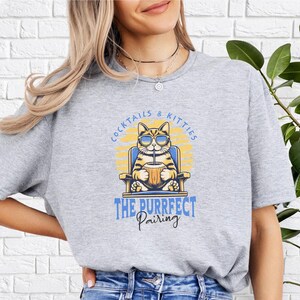 May include: Gray t-shirt with a graphic of a cat sitting in a chair and drinking from a glass. The text on the shirt reads "Cocktails & Kitties The Purrfect Pairing".