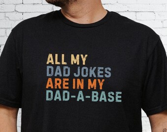 May include: A black short-sleeved t-shirt featuring the text ALL MY DAD JOKES ARE IN MY DAD-A-BASE printed in a distressed, multi-colored font. The words are stacked in yellow, blue, orange, and light green, creating a bold graphic design on the chest.