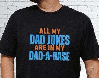 May include: A black short-sleeved t-shirt featuring the orange and blue text ALL MY DAD JOKES ARE IN MY DAD-A-BASE printed on the front. The lettering has a distressed, weathered texture and is centered on the chest of the garment.