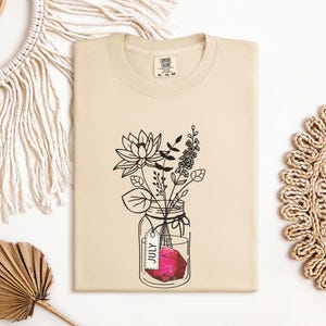 May include: A beige t-shirt featuring a black line drawing of flowers in a mason jar with a tag that says "July". The shirt is made of a soft material and is a neutral color.