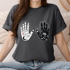 May include: A dark gray t-shirt with a white graphic of two hands. The left hand has a heart and a crescent moon, and the right hand has an eye and a star. The text on the shirt reads "May your light shine from the inside out".