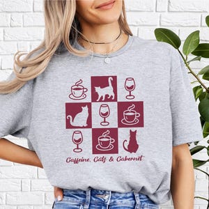 May include: Gray t-shirt with a maroon design featuring a grid of alternating coffee cups, wine glasses, and cats. The text "Caffeine. Cats & Cabernet" is printed below the grid.