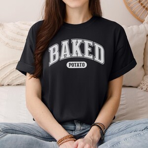 Könnte beinhalten: Schwarzes T-Shirt mit den Worten "BAKED POTATO" in weißer, bogenförmiger Schrift. Das Wort "POTATO" steht in einem kleineren, weißen, ovalen Feld unter "BAKED". Das Shirt hat kurze Ärmel und wird getragen.