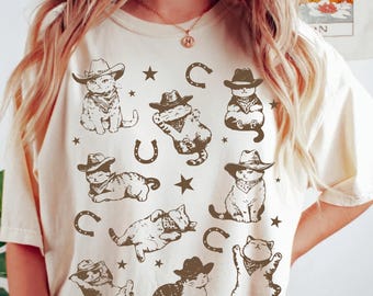 May include: Ivory t-shirt with a brown print of cats wearing cowboy hats and bandanas. The cats are in various poses, including sitting, lying down, and standing. There are also stars and horseshoes printed on the shirt.