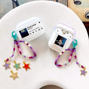 Könnte beinhalten: Zwei weiße AirPod-Hüllen mit transparenten Deckeln, jeweils mit einem Musik-Themen-Design. Eine Hülle zeigt "Wildflower" von Billie Eilish, die andere "Rehab" von Amy Winehouse. Beide Hüllen haben Perlenriemen mit Sternanhängern in verschiedenen Farben.