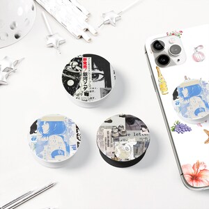 May include: A collection of round phone grips with various designs. One features a manga-style eye and text, another a blue-toned anime illustration, and the third a collage of images and text. A phone with a matching grip is also visible.