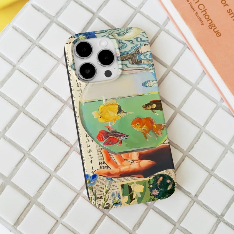 May include: A phone case featuring a colorful fishbowl design with various fish. The case has a collage-style background with floral and Asian-inspired elements. The case is on a white tile surface.