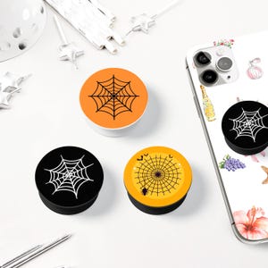 May include: A collection of Halloween-themed phone grips. The grips feature a black spider web design on orange, black, and yellow backgrounds. One grip is attached to a white phone case with floral and perfume bottle designs.