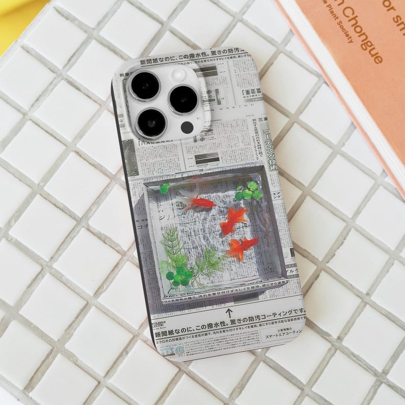 May include: A smartphone case featuring a newspaper print design with a clear rectangular window. Inside the window, there are three orange goldfish swimming amongst green plants. The case has a glossy finish and a camera cutout.