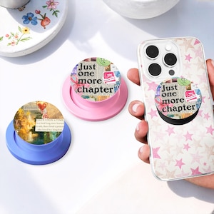 May include: Two circular phone grips and a clear phone case with a pink star pattern. One grip is blue with a fantasy landscape illustration and text. The other grip and the one attached to the phone feature a collage design with the text Just one more chapter.