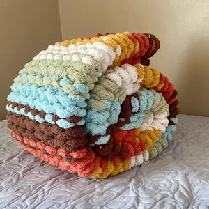 May include: A chunky knit blanket featuring a striped pattern of thick, soft yarn in shades of brown, orange, yellow, white, light green, and light blue. The blanket is folded into a rounded, textured bundle, showcasing the thick, looped yarn texture.