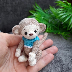 May include: A small crocheted monkey toy with a blue and white shirt. The monkey has brown eyes and a brown nose. The monkey is sitting on a hand.
