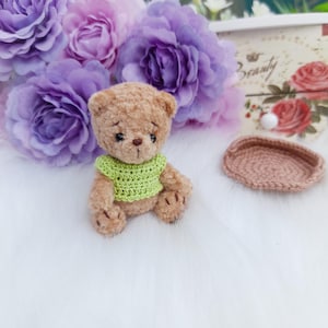 May include: A small brown teddy bear wearing a green crocheted sweater sits on a white fluffy surface. The bear has a sweet expression and is surrounded by purple flowers.