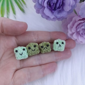 May include: Four small crocheted squares with different facial expressions. The squares are green and light green in color.