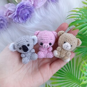 May include: Three small crocheted stuffed animals: a grey koala, a pink pig, and a brown bear. The animals are sitting on a hand.