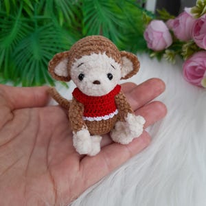 May include: A small brown crocheted monkey wearing a red sweater with white trim. The monkey has white fur on its face and hands.