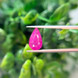 May include: A pear-shaped, faceted gemstone in a vibrant, saturated pink color. The gemstone is held securely between the two metal tips of a pair of silver-toned tweezers, showcasing its clear, geometric cut and brilliant, reflective surface.