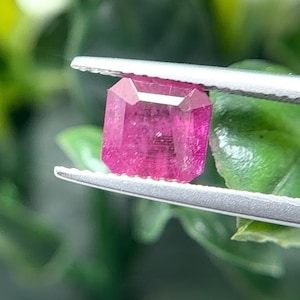 May include: A vibrant, deep pink gemstone with a rectangular step-cut facet pattern. The stone is held securely by a pair of silver-toned metal tweezers, showcasing its translucent quality and sharp, geometric edges against a blurred green background.