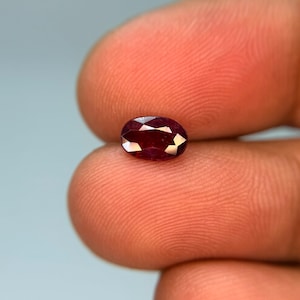 Könnte beinhalten: Ein kleiner, oval geschliffener Edelstein mit einer tiefen, gesättigten roten Farbe und zahlreichen dreieckigen Facetten, die das Licht reflektieren. Der Stein weist eine hohe Reinheit und eine polierte Oberfläche auf, die einen brillanten Farbton zeigt.
