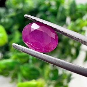May include: A vibrant, oval-cut magenta gemstone held by a pair of silver-toned metal tweezers. The stone features a faceted surface that reflects light, showcasing a deep pinkish-red hue with a polished, translucent appearance and sharp edges.