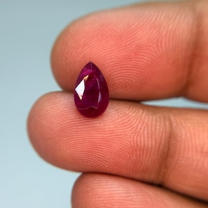 Puede incluir: Una piedra preciosa de rubí tallada en forma de pera con un color rojo profundo y saturado. La piedra presenta una parte superior puntiaguda y una base redondeada, mostrando múltiples facetas geométricas que reflejan la luz sobre su superficie pulida.