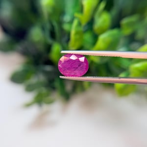 May include: A vibrant, oval-cut magenta gemstone with a faceted surface held by silver-toned metal tweezers. The stone displays internal inclusions and a deep pink hue, showcasing a polished finish that reflects light across its multiple facets.