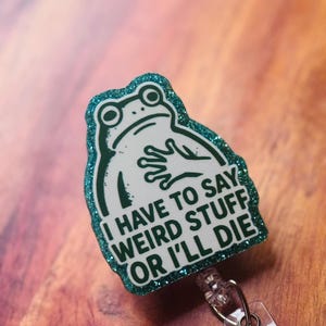 May include: A badge reel with a green glitter border and a white frog illustration. The text reads "I HAVE TO SAY WEIRD STUFF OR I'LL DIE". The reel has a clear plastic strap and a metal clip.