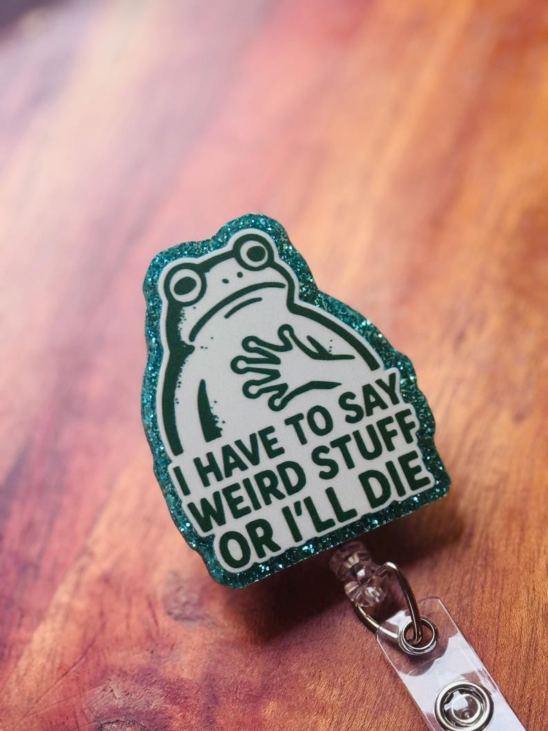 May include: A badge reel with a green glitter border and a white frog illustration. The text reads "I HAVE TO SAY WEIRD STUFF OR I'LL DIE". The reel has a clear plastic strap and a metal clip.