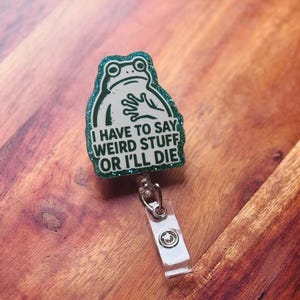 May include: A green glitter badge reel with a frog design. The frog has a surprised expression and is holding its hands up. The text reads, "I HAVE TO SAY WEIRD STUFF OR I'LL DIE."