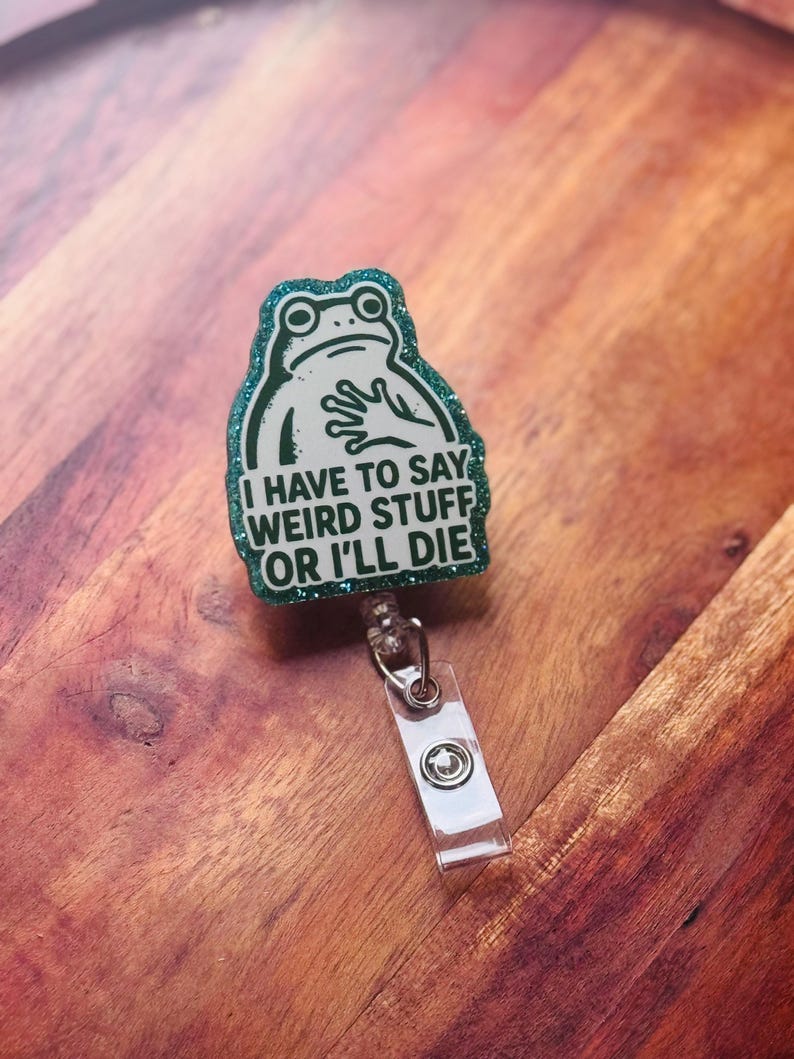 May include: A green glitter badge reel with a frog design. The frog has a surprised expression and is holding its hands up. The text reads, "I HAVE TO SAY WEIRD STUFF OR I'LL DIE."
