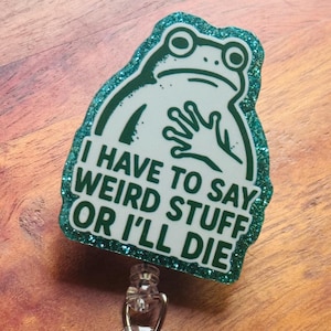 May include: A badge reel with a green glitter border and a white frog illustration. The text "I HAVE TO SAY WEIRD STUFF OR I'LL DIE" is printed on the badge. The reel has a clear plastic strap and a metal clip.
