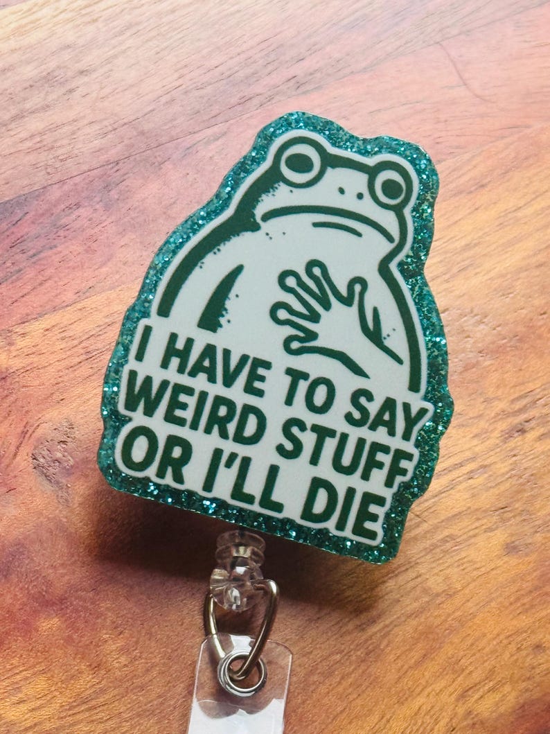 May include: A badge reel with a green glitter border and a white frog illustration. The text "I HAVE TO SAY WEIRD STUFF OR I'LL DIE" is printed on the badge. The reel has a clear plastic strap and a metal clip.