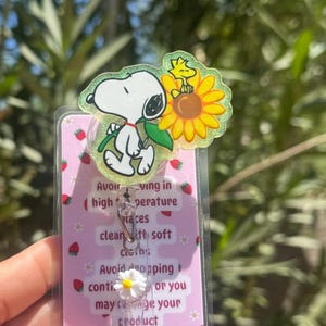 May include: A Snoopy-themed badge reel with a clear plastic cover featuring Snoopy, Woodstock, and a sunflower. The badge reel has a pink background with strawberry and flower designs, and text that reads "Avoid..." and "product."