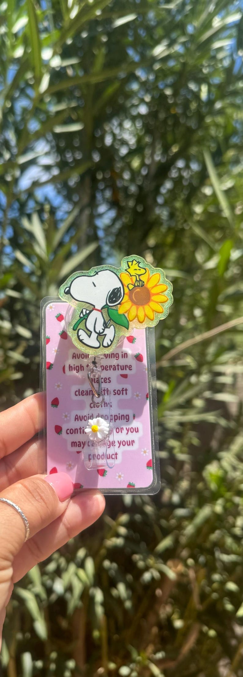 May include: A Snoopy-themed badge reel with a clear plastic cover featuring Snoopy, Woodstock, and a sunflower. The badge reel has a pink background with strawberry and flower designs, and text that reads "Avoid..." and "product."