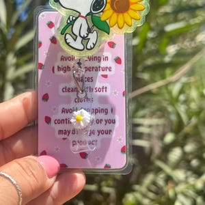May include: A Snoopy-themed retractable badge holder with a daisy charm. The badge reel features Snoopy with a sunflower and the text "Avoiding in (high temperature) laces clean with soft cloth: Avoid dropping or you may damage your product."