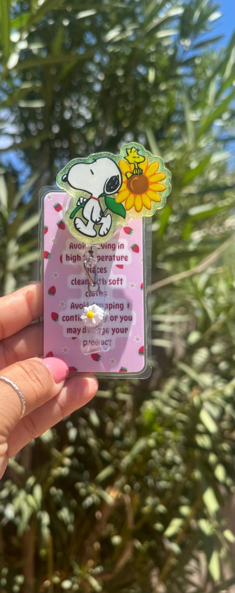 May include: A Snoopy-themed retractable badge holder with a daisy charm. The badge reel features Snoopy with a sunflower and the text "Avoiding in (high temperature) laces clean with soft cloth: Avoid dropping or you may damage your product."