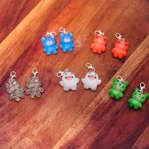 May include: A collection of colorful, small monster-shaped charms. The charms are in various colors, including blue, orange, white, green, and leopard print. Each charm has a silver-colored clasp attached. The charms are arranged on a wooden surface.