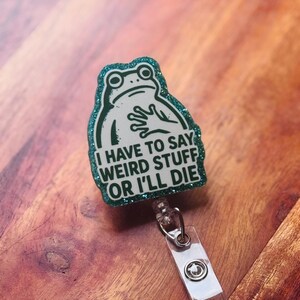 May include: A badge reel with a green glitter border and a white frog illustration. The text on the badge reel reads "I HAVE TO SAY WEIRD STUFF OR I'LL DIE". The reel has a clear plastic strap and a metal clip.