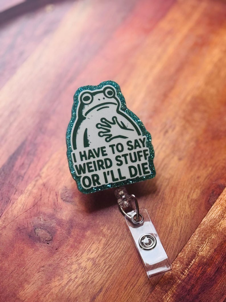 May include: A badge reel with a green glitter border and a white frog illustration. The text on the badge reel reads "I HAVE TO SAY WEIRD STUFF OR I'LL DIE". The reel has a clear plastic strap and a metal clip.