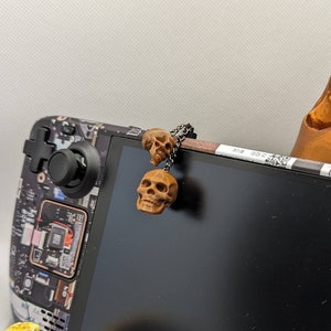 May include: A black and white gaming console with a skull charm hanging from the top. The console has a black screen and a skull design on the back. The console is sitting on a white surface.